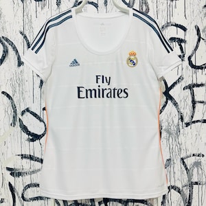 May include: White Adidas soccer jersey with navy blue stripes on the shoulders and the text "Fly Emirates". The jersey features the Adidas logo and a team crest. Orange accents are visible on the sides.