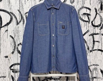 May include: A blue denim shirt with two chest pockets and a button-down front. The shirt has a small label on the right chest pocket. The shirt is hanging on a white hanger against a graffiti-style background.