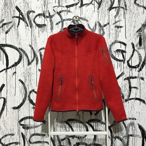 Puede incluir: Una chaqueta de forro polar roja con cierre de cremallera y varios bolsillos. La chaqueta tiene un cuello alto y un forro morado oscuro.