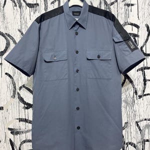 May include: A short-sleeved, slate blue button-up shirt with black shoulder accents and two chest pockets. The shirt features the Versace logo on the label and sleeve. The shirt is hanging on a white hanger.