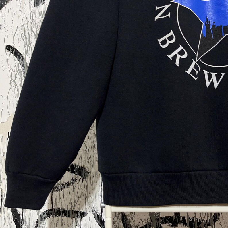 May include: Black sweatshirt with the text "N BREW" in white, arched around a blue graphic. The graphic appears to be a silhouette of a city skyline. The sweatshirt has a ribbed hem and cuffs.