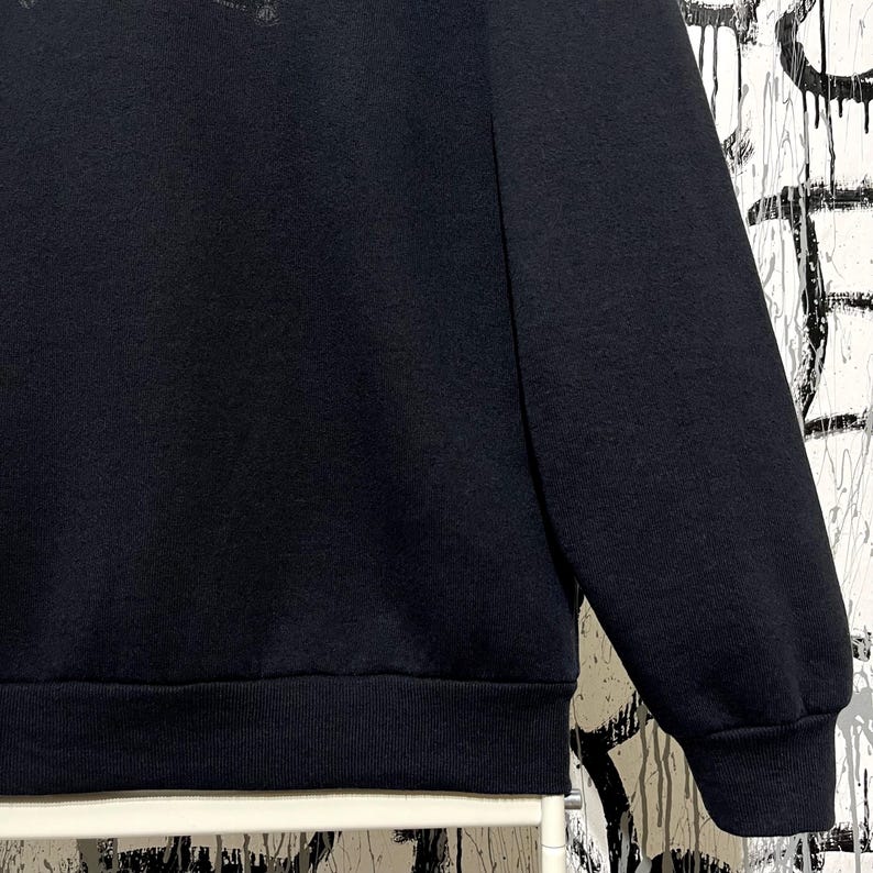 May include: A close-up of a black sweatshirt. The image shows the sleeve and part of the body of the garment. The fabric appears to be a soft, textured material. The cuff and hem are ribbed.