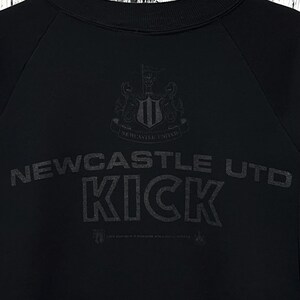 May include: Black sweatshirt featuring the Newcastle United crest and the words "Newcastle Utd KICK" in a dark grey font. The sweatshirt has a crew neck and long sleeves. The design is printed on the front.