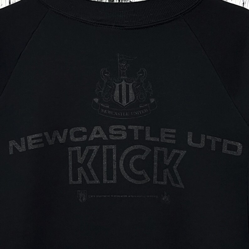 May include: Black sweatshirt featuring the Newcastle United crest and the words "Newcastle Utd KICK" in a dark grey font. The sweatshirt has a crew neck and long sleeves. The design is printed on the front.