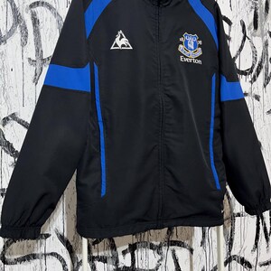 K&ouml;nnte beinhalten: Schwarze und blaue Everton-Fu&szlig;balljacke. Die Jacke hat blaue Eins&auml;tze an Schultern und &Auml;rmeln, mit dem Everton-Vereinswappen und Text auf der Brust. Sie hat einen durchgehenden Rei&szlig;verschluss und einen hohen Kragen.