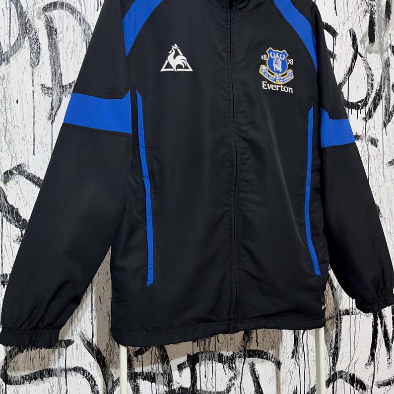 K&ouml;nnte beinhalten: Schwarze und blaue Everton-Fu&szlig;balljacke. Die Jacke hat blaue Eins&auml;tze an Schultern und &Auml;rmeln, mit dem Everton-Vereinswappen und Text auf der Brust. Sie hat einen durchgehenden Rei&szlig;verschluss und einen hohen Kragen.