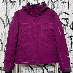 Puede incluir: Una chaqueta de invierno Columbia magenta con capucha. La chaqueta tiene múltiples bolsillos, cierre de cremallera y la palabra "Titanium" impresa en la manga. Los puños son negros con ribete gris. La chaqueta está colgada en una percha blanca.