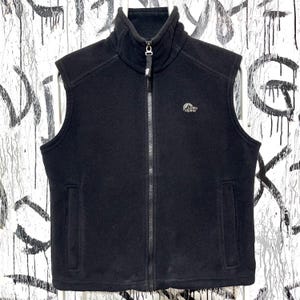 May include: Black fleece vest with a high collar and a full-length zipper. The vest has two front pockets and the "Lowe Alpine" logo on the chest. The vest is sleeveless and suitable for outdoor activities.