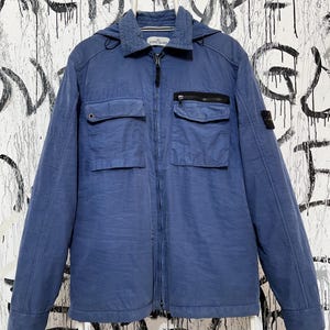 May include: A blue hooded jacket with two chest pockets and two lower pockets. It features a zip closure and a small logo patch on the left sleeve. The jacket is suspended from a white hanger.