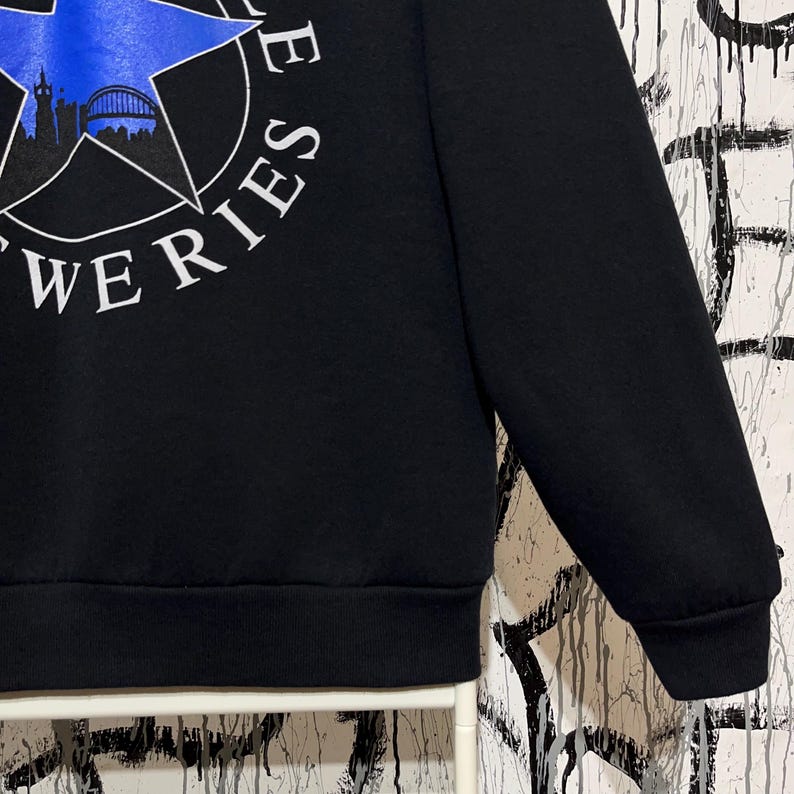 May include: Black sweatshirt with a white and blue star graphic. The graphic includes the text "WERIES" and a silhouette of a bridge and buildings. The sweatshirt has a crew neck and long sleeves.