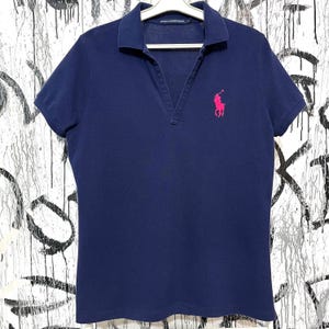 May include: A navy blue polo shirt with short sleeves and a deep V-neck. The shirt features a pink embroidered logo of a polo player on the left chest. The shirt is hanging on a white hanger.