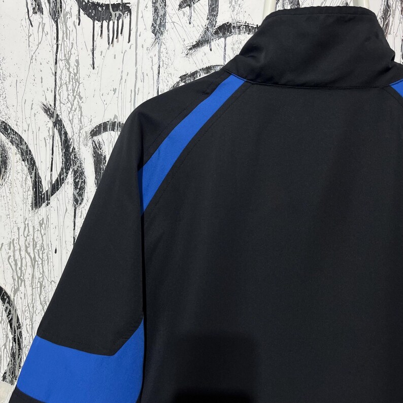 K&ouml;nnte beinhalten: Schwarze Jacke mit blauen Schulter- und &Auml;rmelakzenten. Die Jacke hat einen hohen Kragen und einen durchgehenden Rei&szlig;verschluss. Das Material scheint glatt, m&ouml;glicherweise wasserabweisend. Der Hintergrund ist eine wei&szlig;e Wand mit schwarzen Farbspritzern.