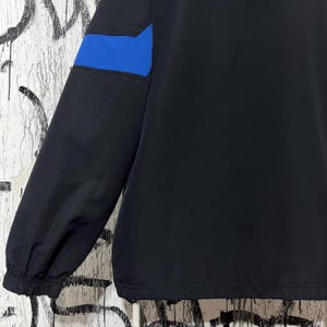 K&ouml;nnte beinhalten: Schwarze Jacke mit blauem Akzent am oberen &Auml;rmel. Die Jacke hat ein schlichtes Design mit geripptem B&uuml;ndchen und glatter, matter Oberfl&auml;che. Der blaue Akzent setzt einen Farbakzent.