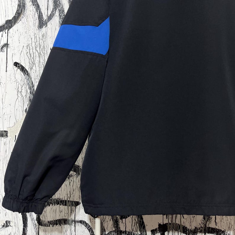 K&ouml;nnte beinhalten: Schwarze Jacke mit blauem Akzent am oberen &Auml;rmel. Die Jacke hat ein schlichtes Design mit geripptem B&uuml;ndchen und glatter, matter Oberfl&auml;che. Der blaue Akzent setzt einen Farbakzent.
