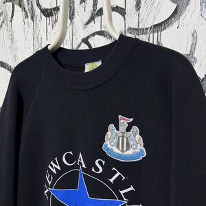 May include: Black crewneck sweatshirt featuring the Newcastle United crest and team name. The crest includes a shield, two seahorses, and a flag. The team name is printed in white with a blue star.