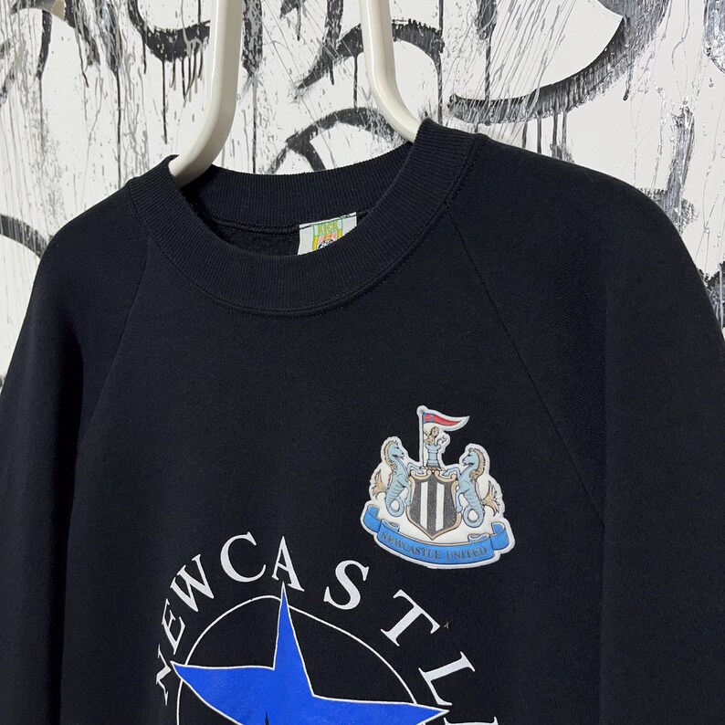 May include: Black crewneck sweatshirt featuring the Newcastle United crest and team name. The crest includes a shield, two seahorses, and a flag. The team name is printed in white with a blue star.
