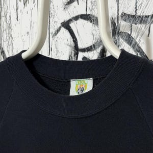 May include: A close-up of a black sweatshirt with a crew neck. The sweatshirt has a tag with the word "KICK" and a globe design. The garment is hanging on a white hanger against a background with graffiti.
