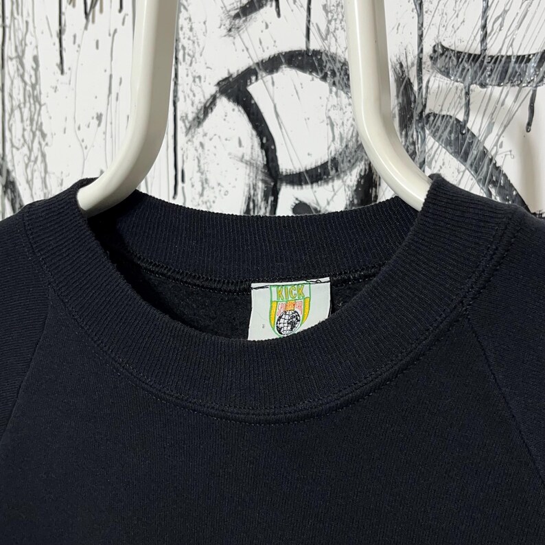 May include: A close-up of a black sweatshirt with a crew neck. The sweatshirt has a tag with the word "KICK" and a globe design. The garment is hanging on a white hanger against a background with graffiti.