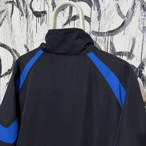K&ouml;nnte beinhalten: Schwarze Jacke mit hohem Kragen und blauen Streifen auf den Schultern. Die Jacke hat ein sportliches Design und besteht aus einem glatten Material. Die R&uuml;ckseite der Jacke ist sichtbar und zeigt den Farbkontrast.