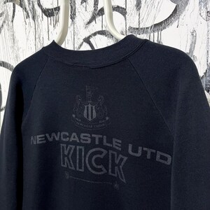 May include: Black sweatshirt featuring the text "Newcastle Utd KICK" in a distressed font. The Newcastle United crest is above the text. The sweatshirt has a crew neck and raglan sleeves.