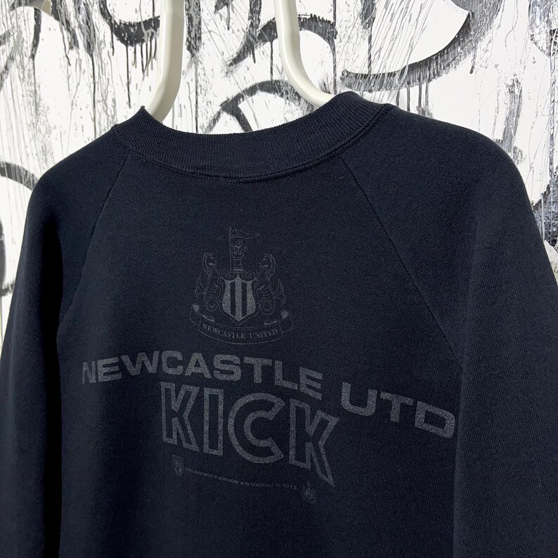 May include: Black sweatshirt featuring the text "Newcastle Utd KICK" in a distressed font. The Newcastle United crest is above the text. The sweatshirt has a crew neck and raglan sleeves.