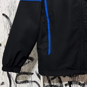 K&ouml;nnte beinhalten: Schwarze Jacke mit blauen Akzenten. Die Jacke hat einen blauen Streifen am &Auml;rmel und &uuml;ber dem oberen R&uuml;cken. Sie hat einen Rei&szlig;verschluss und einen Stehkragen. Die Jacke ist aus leichtem Material gefertigt.