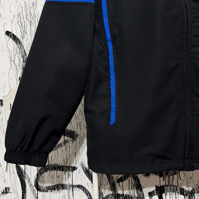 K&ouml;nnte beinhalten: Schwarze Jacke mit blauen Akzenten. Die Jacke hat einen blauen Streifen am &Auml;rmel und &uuml;ber dem oberen R&uuml;cken. Sie hat einen Rei&szlig;verschluss und einen Stehkragen. Die Jacke ist aus leichtem Material gefertigt.