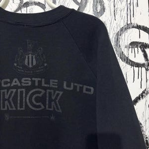 May include: Black sweatshirt with the text "CASTLE UTD KICK" printed in gray. The Newcastle United crest is above the text. The sweatshirt has a crew neck and raglan sleeves.
