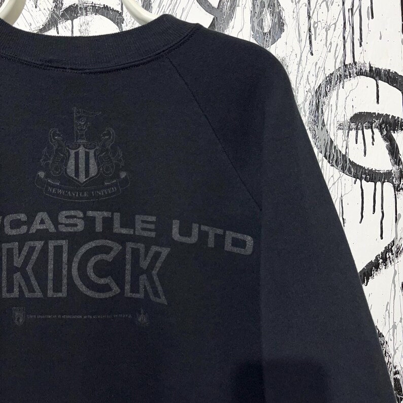 May include: Black sweatshirt with the text "CASTLE UTD KICK" printed in gray. The Newcastle United crest is above the text. The sweatshirt has a crew neck and raglan sleeves.