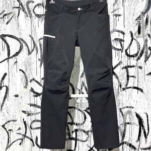 Könnte beinhalten: Schwarze Outdoor-Hose mit Knopfverschluss, Gürtelschlaufen und mehreren Taschen, darunter eine mit weißem Reißverschluss. Die Hose hängt vor einer weißen Wand mit schwarzen Graffiti.