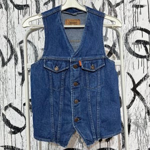 Può includere: Un gilet in denim vintage blu medio, con scollo a V e chiusura con bottoni. Ha due tasche frontali e una piccola etichetta vicino allo scollo. Il gilet è appeso a una gruccia bianca.