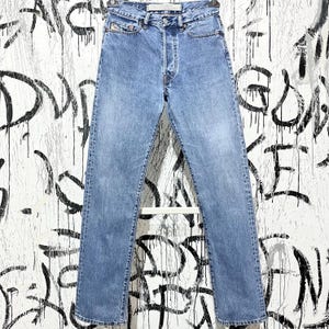 May include: Pale blue denim jeans with a traditional five-pocket design. The jeans feature a button fly and a straight leg. The jeans are hung on a white hanger against a graffiti-covered wall.