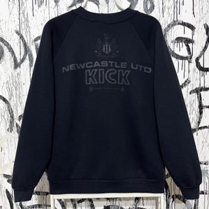 May include: Black sweatshirt with the text "NEWCASTLE UTD KICK" in gray. The sweatshirt has a crew neck and long sleeves. The Newcastle United crest is above the text.