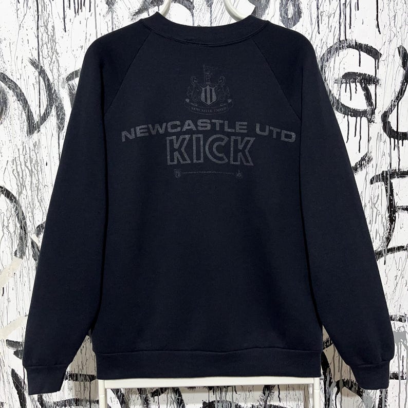 May include: Black sweatshirt with the text "NEWCASTLE UTD KICK" in gray. The sweatshirt has a crew neck and long sleeves. The Newcastle United crest is above the text.