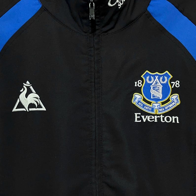 K&ouml;nnte beinhalten: Schwarz-blaue Rei&szlig;verschlussjacke mit dem Everton-Teamwappen auf der Brust. Das Wappen zeigt einen Schild mit einem Turm, die Zahlen 18 und 78 und die Worte "Nil Satis Nisi Optimum". Auf der linken Seite befindet sich ein wei&szlig;es Hahn-Logo.