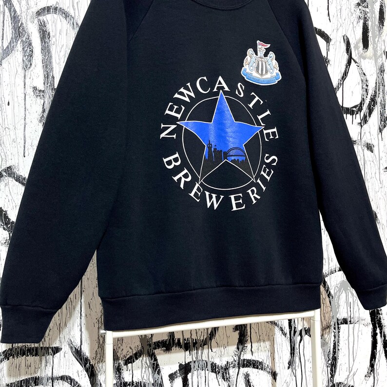May include: Black sweatshirt with the text "NEWCASTLE BREWERIES" in white around a blue star. The star contains a silhouette of a bridge and buildings. A crest is above the star. The sweatshirt is a raglan style.