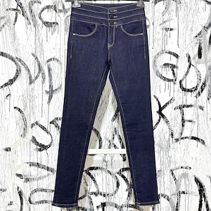 May include: Dark wash denim jeans with a high waist and three button closure. The jeans have classic five-pocket styling and a slim fit. The jeans are displayed on a white hanger against a graffiti-covered wall.