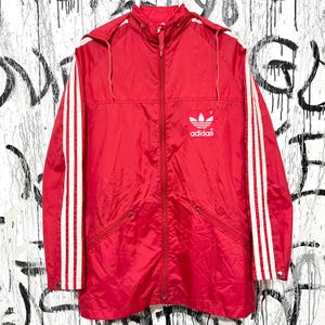 Op de afbeelding: Een rode Adidas jas met een capuchon en witte strepen langs de mouwen. De jas heeft een ritssluiting en het Adidas logo op de borst. De jas is gemaakt van een lichtgewicht materiaal.