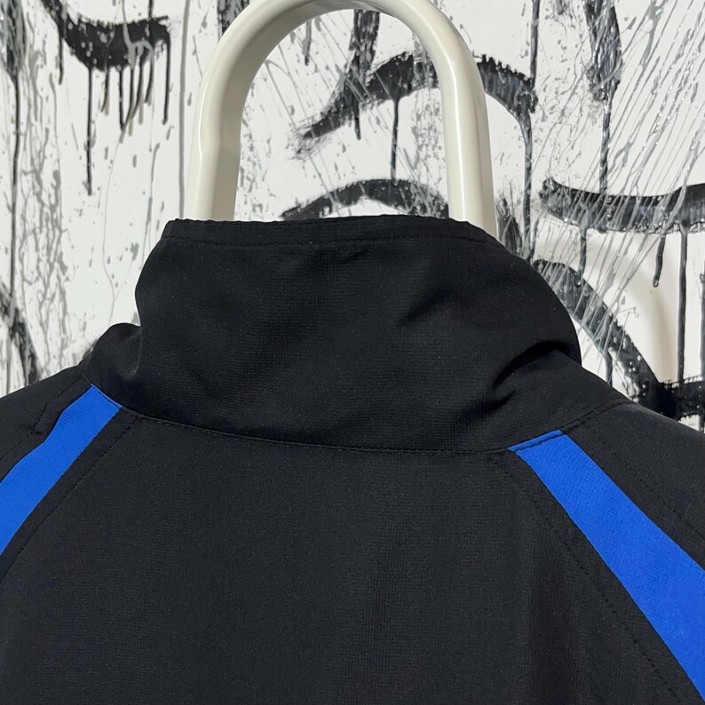 K&ouml;nnte beinhalten: Schwarze Jacke mit hohem Kragen und blauen Streifen an den &Auml;rmeln. Die Jacke h&auml;ngt an einem wei&szlig;en Kleiderb&uuml;gel vor einem Hintergrund mit schwarzen und wei&szlig;en Farbspritzern.