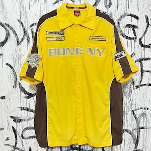 Puede incluir: Camisa amarilla con botones con detalles en marrón y ribete blanco. La camisa presenta el texto bordado "BONE NY" y parches que dicen "peedway", "WRS 2004" y "champion series". La camisa de manga corta tiene cuello.