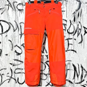 Könnte beinhalten: Leuchtend orangefarbene Schneehose mit mehreren Taschen und Reißverschlüssen. Die Hose hat einen verstärkten Kniebereich und das Peak Performance-Logo. Die Hose hängt an einem schwarzen Kleiderbügel vor einem Graffiti-Hintergrund.