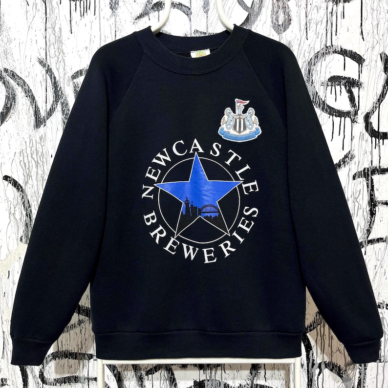May include: Black crew neck sweatshirt with a blue star graphic and the words "Newcastle Breweries" in white. The sweatshirt also features a crest with a shield and the words "Newcastle United".