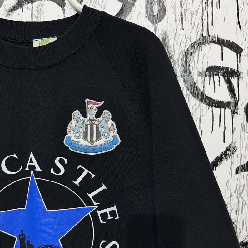 May include: Black sweatshirt featuring a Newcastle United crest and logo. The crest includes two seahorses, a shield, and a red flag. The word "CASTLES" is visible, along with a blue star and a silhouette of a castle.