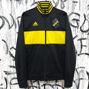 May include: Black Adidas track jacket with a yellow horizontal stripe across the chest. The jacket has a yellow collar and the Adidas logo on the chest. A patch with text is on the right chest.