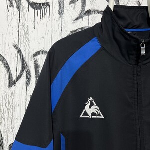 K&ouml;nnte beinhalten: Schwarze Sportjacke mit blauen Streifen auf den &Auml;rmeln. Verf&uuml;gt &uuml;ber ein wei&szlig;es Logo eines Hahns in einem Dreieck. Die Jacke hat einen durchgehenden Rei&szlig;verschluss und einen hohen Kragen.