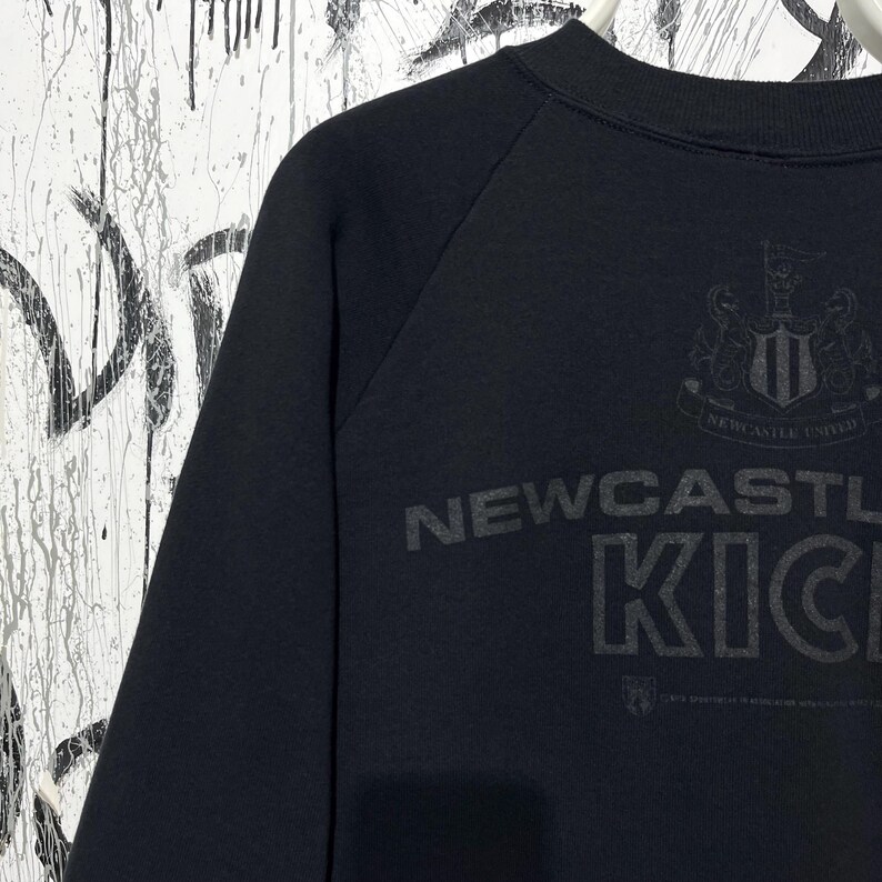 May include: Black sweatshirt with the text "NEWCASTLE KICKS" in a dark grey font. The Newcastle United crest is visible above the text. The sweatshirt has raglan sleeves and a crew neck.