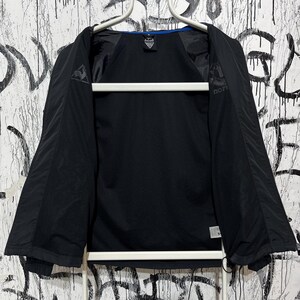 K&ouml;nnte beinhalten: Schwarze Sportjacke mit blauem Besatz am Kragen. Die Jacke ist offen und zeigt das Innenfutter. Das Le Coq Sportif-Logo ist auf der Brust und am &Auml;rmel zu sehen. Die Jacke h&auml;ngt an einem wei&szlig;en Kleiderb&uuml;gel.