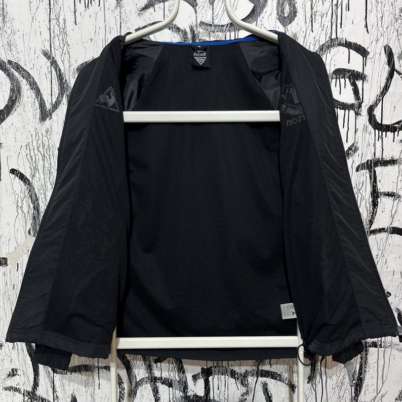 K&ouml;nnte beinhalten: Schwarze Sportjacke mit blauem Besatz am Kragen. Die Jacke ist offen und zeigt das Innenfutter. Das Le Coq Sportif-Logo ist auf der Brust und am &Auml;rmel zu sehen. Die Jacke h&auml;ngt an einem wei&szlig;en Kleiderb&uuml;gel.
