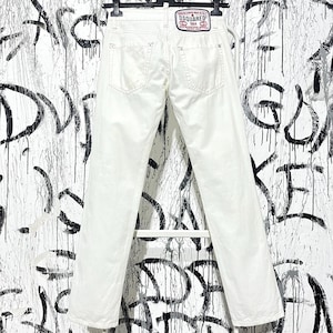 May include: White denim jeans with a straight leg cut, hanging on a black hanger. The back of the jeans features two pockets and a branded patch. The background is a white wall with black graffiti.