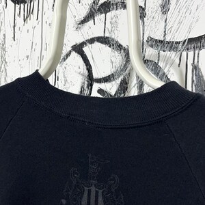 May include: Black crew neck sweatshirt with a tonal emblem on the lower front. The sweatshirt is hanging on a white hanger against a backdrop of black and white abstract art. The emblem appears to be a coat of arms.